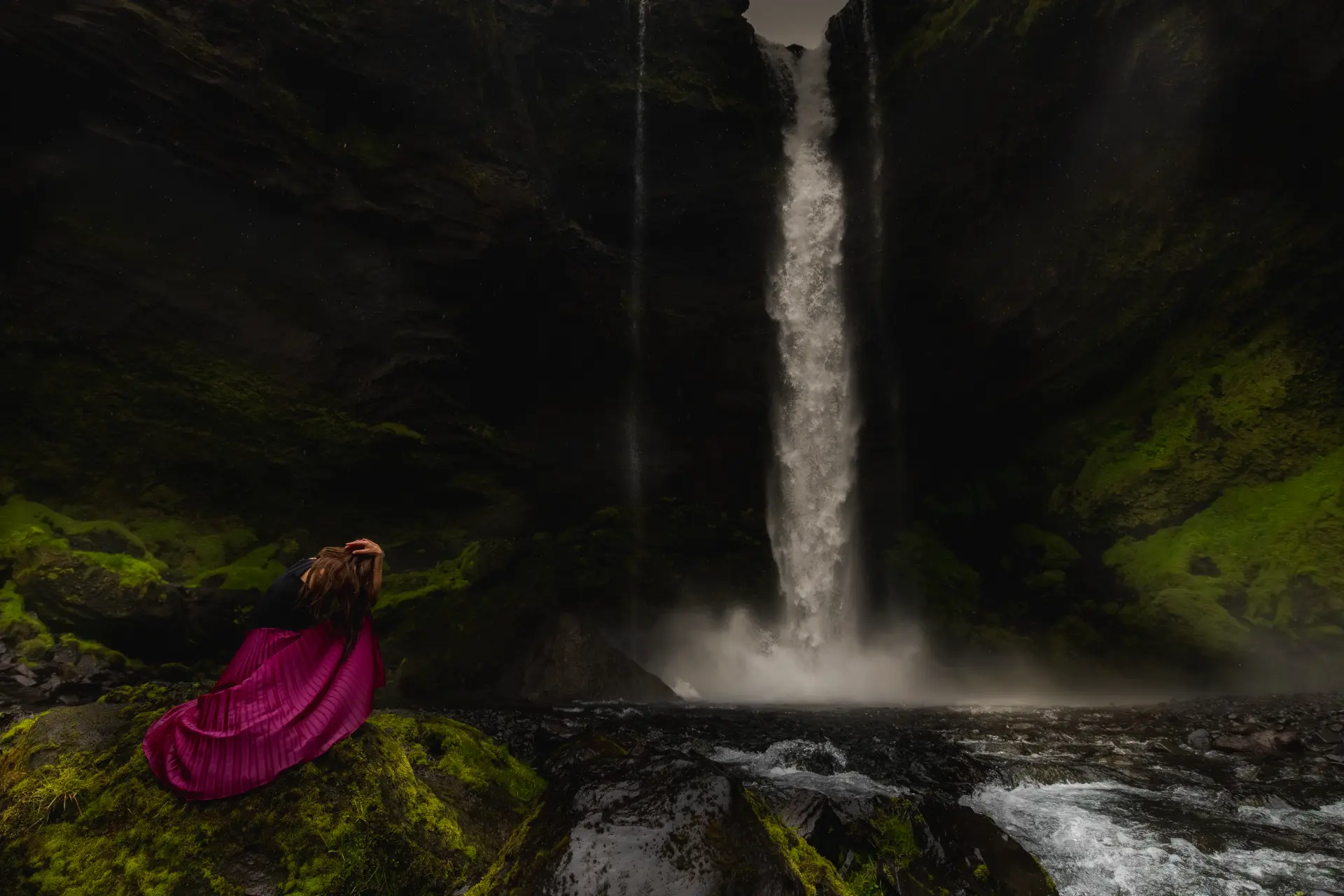 Woman in a magenta pleated skirt beside a moss-walled Icelandic waterfall